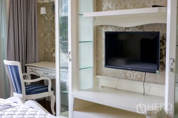 คอนโด ให้เช่าในKhlong Toei, Bangkok - มุมห้องนอนพร้อมตู้ชั้นวางสีขาว ทีวีติดผนัง และโต๊ะทำงานบนวอลเปเปอร์ลายคลาสสิก