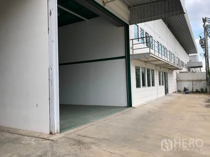Factory for rent in Lam Luk Ka, Pathum Thani - Wide concrete driveway and loading area beside the factory with roll‑up door for trucks.