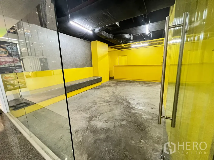 พื้นที่ขายปลีก ให้เช่า - มุมมองจากทางเข้าผ่านประตูกระจกสู่พื้นที่พื้นคอนกรีต ผนังสีเหลืองเด่น