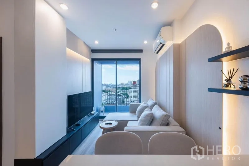 Condominium for rent in Bang Na, Bangkok - Cozy lounge facing the balcony with wall-mounted TV unit, shelves, and sliding glass doors framing the skyline.