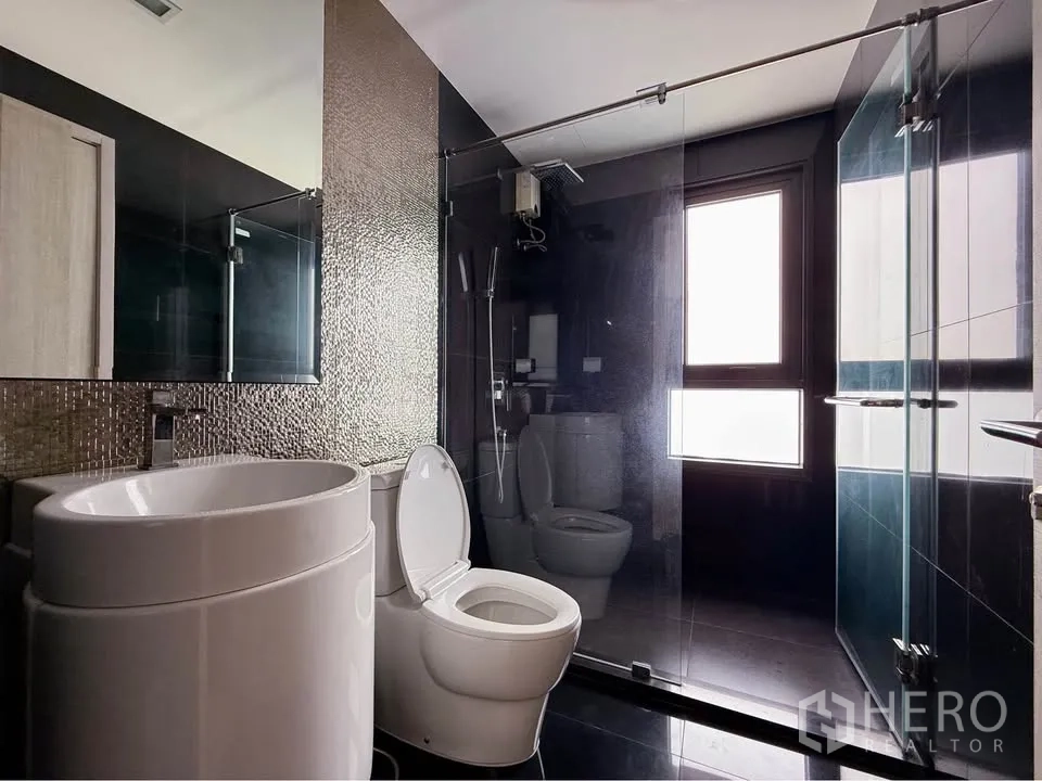 Condominium for rent in Khlong Toei, Bangkok - Modern bathroom with mosaic accent wall, round basin, toilet and glass-enclosed shower near a frosted window.