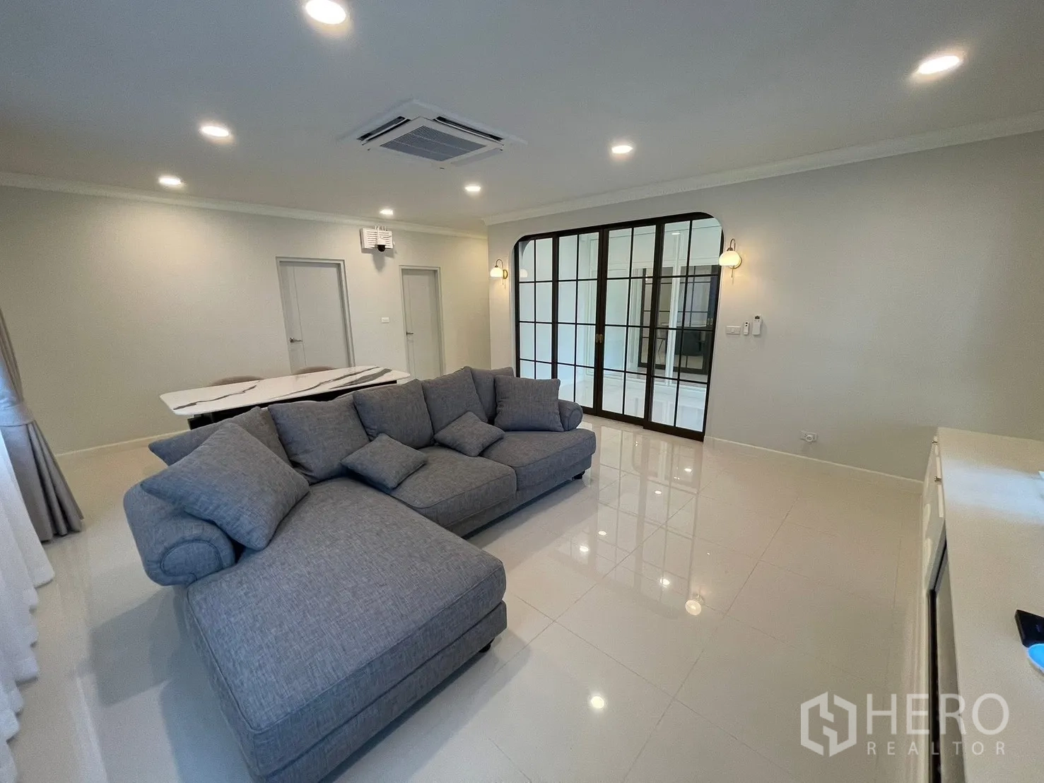 Detached House for rent in Bang Phli, Samut Prakan - Spacious living room with L-shaped sofa, glossy tile floors, recessed lights, and black-framed glass partition doors.