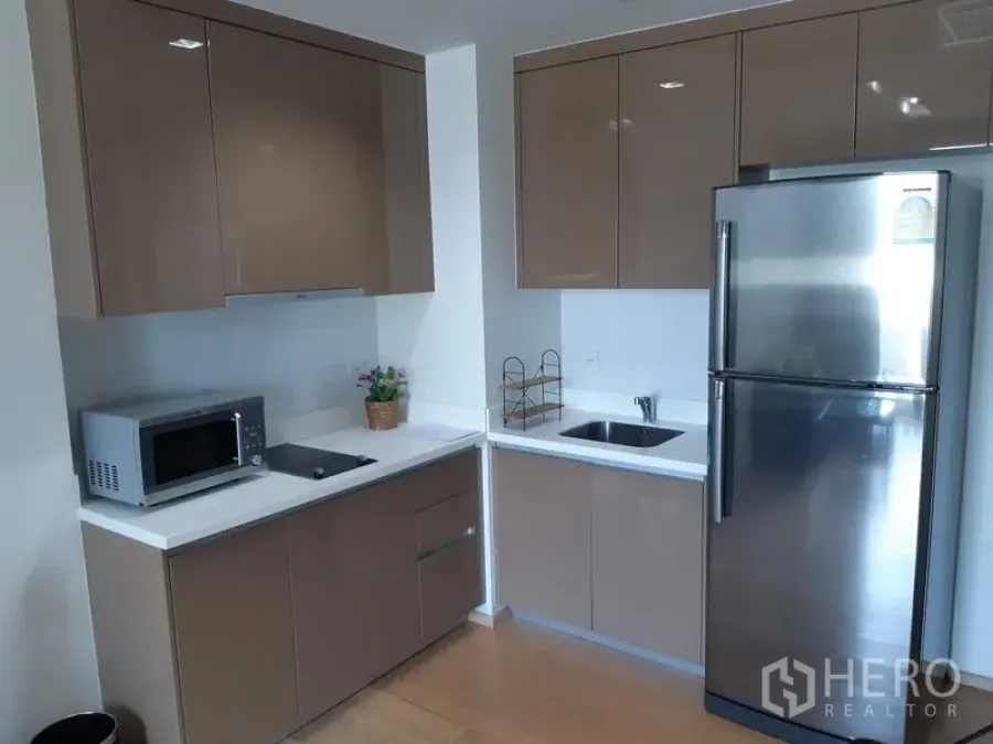 คอนโด ให้เช่าในKhlong Toei, Bangkok - ครัวมุมรูปตัวแอล ตู้บานเงาสีน้ำตาล เคาน์เตอร์ขาว พร้อมตู้เย็นสแตนเลสและไมโครเวฟ