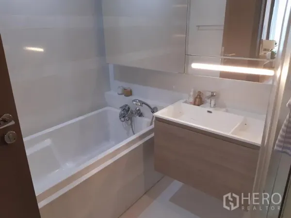 Condominium for rent in Khlong Toei, Bangkok - Second bathroom with white bathtub, wall-mounted vanity, backlit mirror, and neutral tile finishes.
