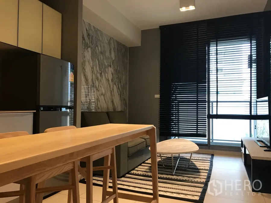 Condominium for rent in Watthana, Bangkok - Modern living room with marble feature wall, wooden dining bar, and floor-to-ceiling windows with black blinds.