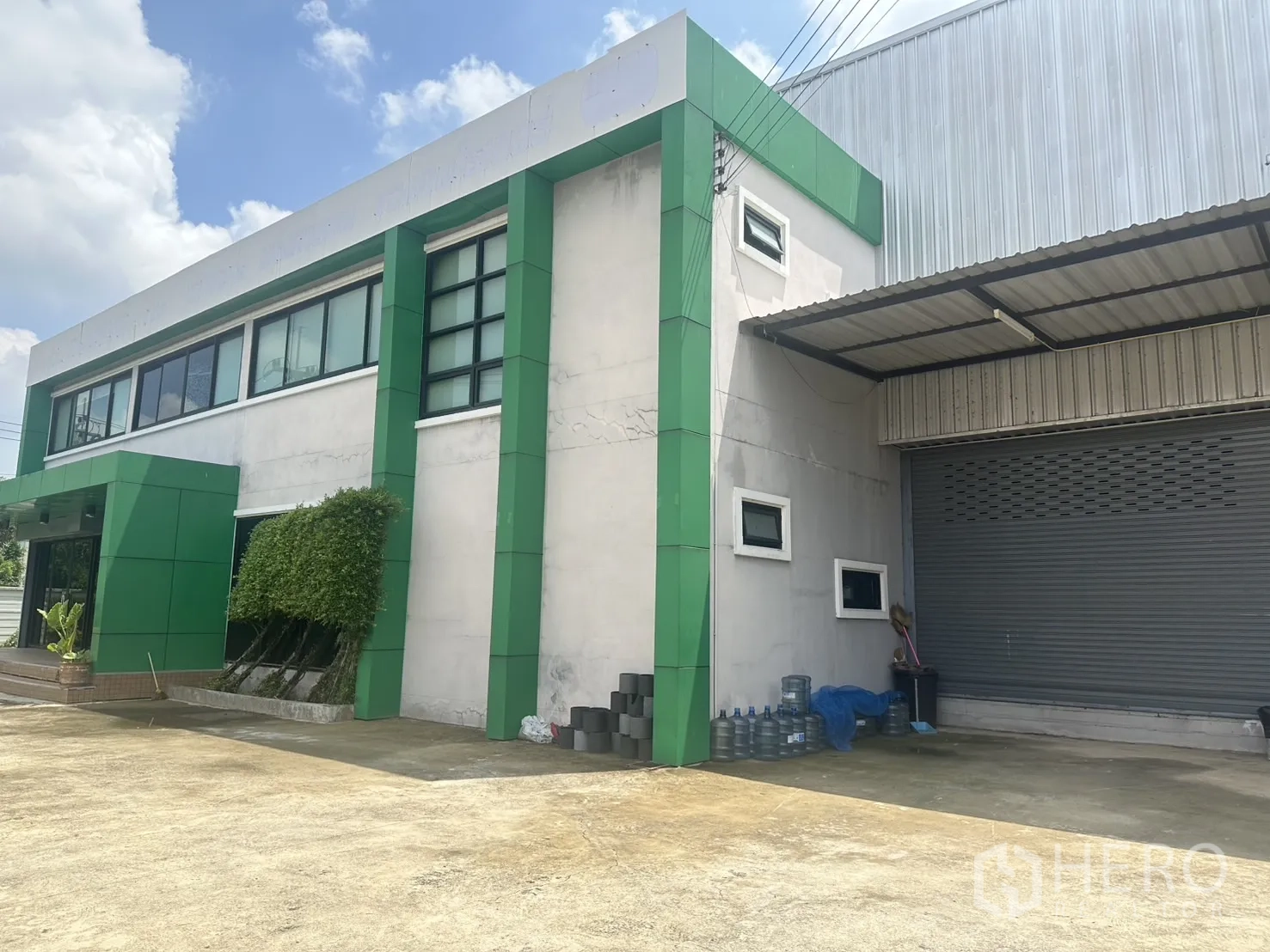 Warehouse for sale in Bang Len, Nakhon Pathom - Close angle of the office facade with shrubs and an adjacent shuttered loading door.