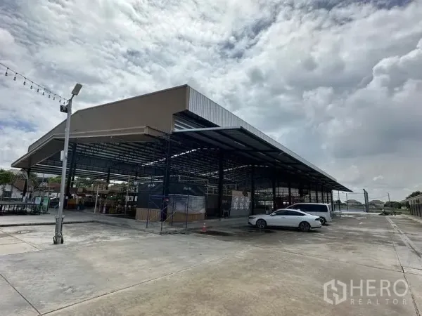 Warehouse for rent in Sai Noi, Nonthaburi - Open-sided steel warehouse with high roof and wide concrete yard with parking and loading area under cloudy sky in Sai Noi, Nonthaburi.