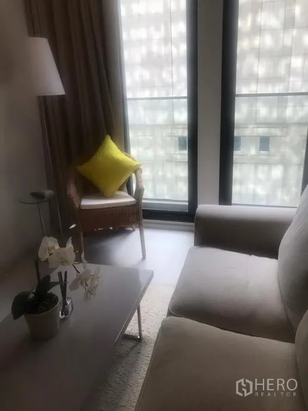 Condominium for rent in Pathum Wan, Bangkok - Cozy living room with sofa, coffee table, floor lamp, and large city-facing window bringing in natural light.