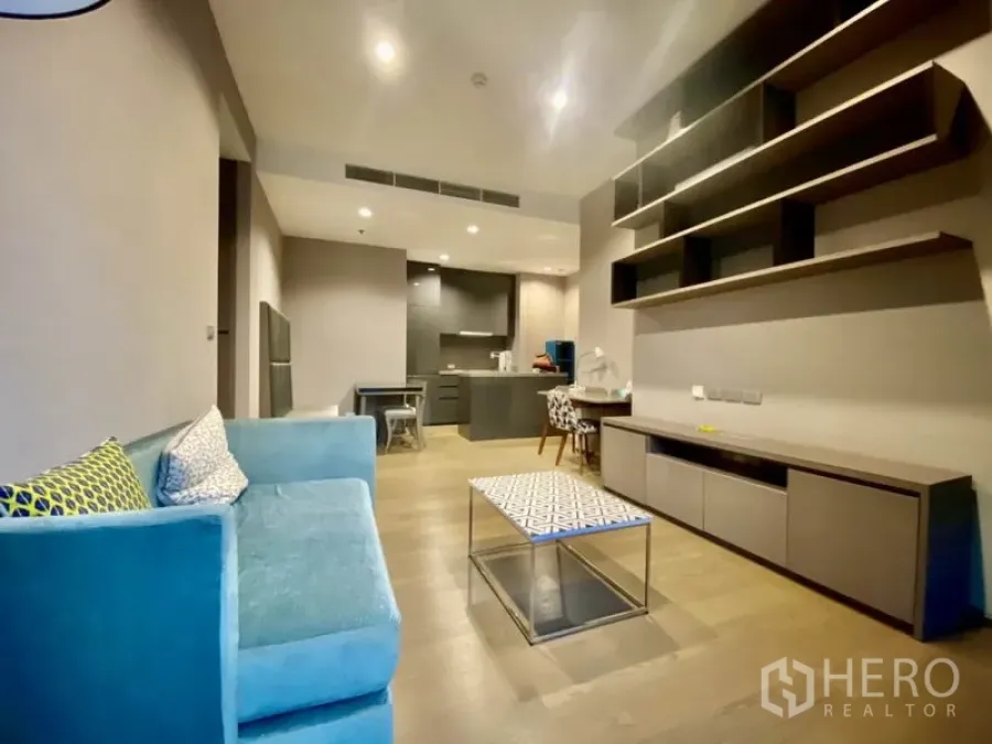 Condominium for rent or sale in Bang Rak, Bangkok - Modern Bang Rak condo living room with teal sofa, wall shelving, TV console and open-plan kitchen on wood floors.