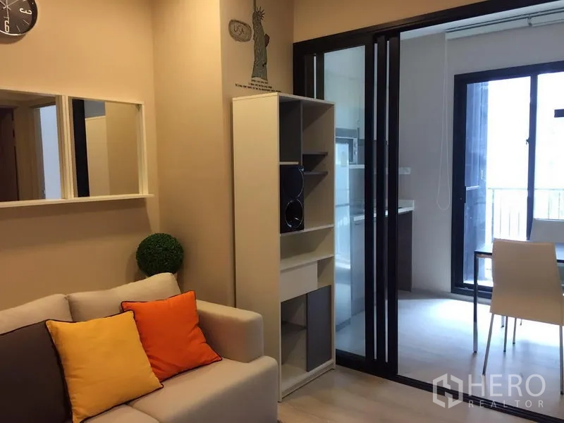 Condominium for sale in Huai Khwang, Bangkok - Cozy living room with sofa and shelving, opening to a kitchenette and balcony via sliding glass doors.