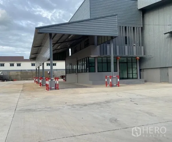 คลังสินค้า ให้เช่าในBang Phli, Samut Prakan - มุมข้างของหลังคาคลุมลานและหน้าต่างสำนักงาน พร้อมพื้นที่ให้รถบรรทุกเลี้ยวเข้าออกสะดวก มีเสากันชนแดง‑ขาว