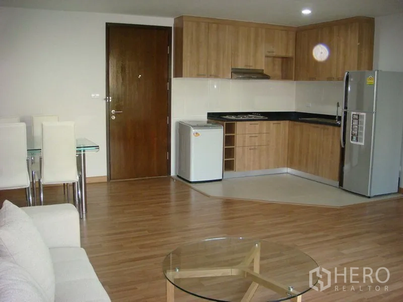 Apartment for rent in Khlong Toei, Bangkok - Open-plan living area with wood-tone L-shaped kitchen, refrigerator and dining set in an Asok apartment.