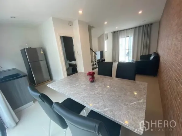 Townhouse for rent or sale in Bang Kapi, Bangkok - Open-plan dining and living area with a marble table, brick feature wall, modern pantry kitchen and staircase, filled with natural light.
