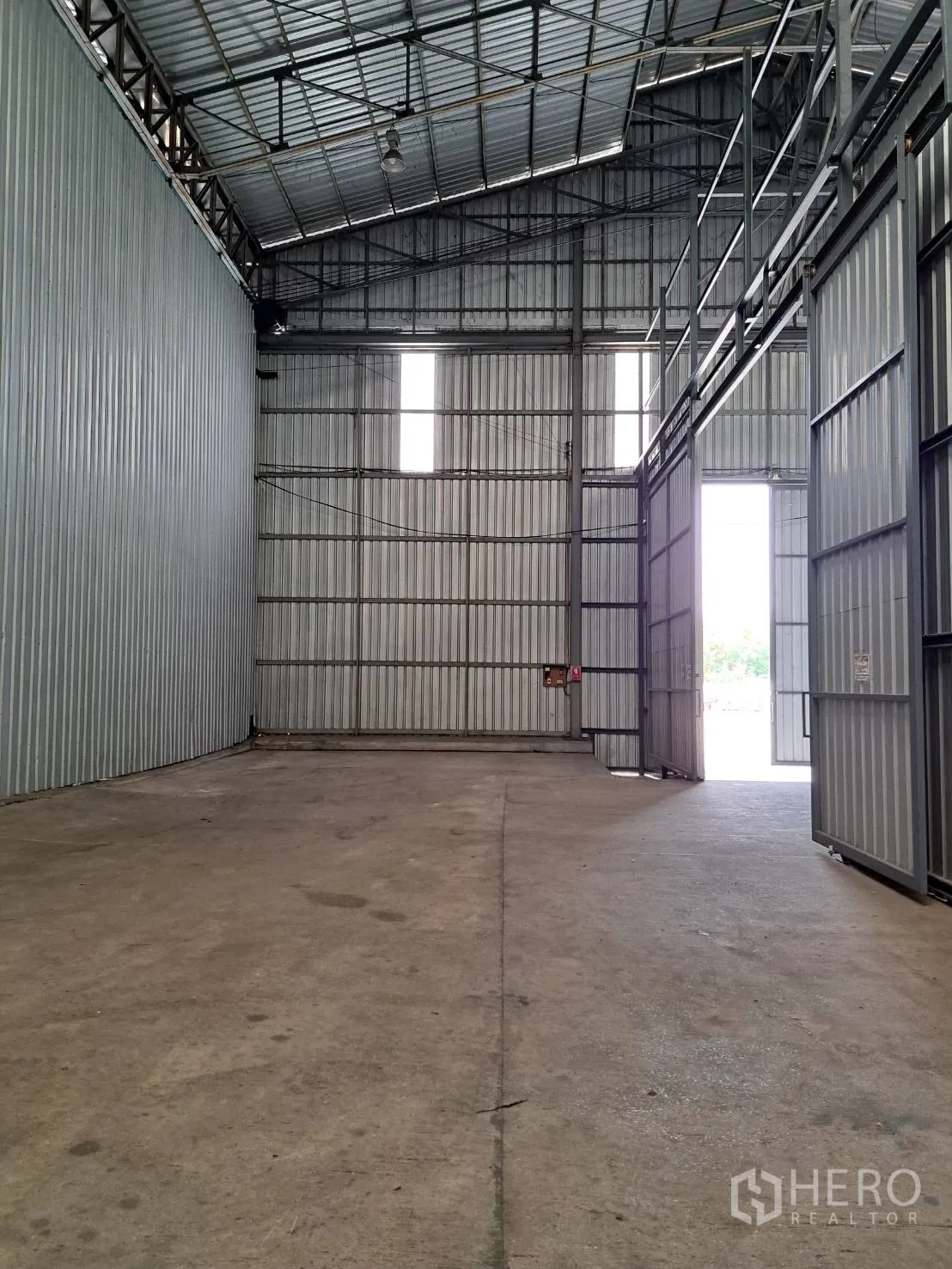 Warehouse for rent in Pak Kret, Nonthaburi - Wide sliding steel doors and tall windows inside an industrial warehouse with open-plan concrete floor and gabled roof.