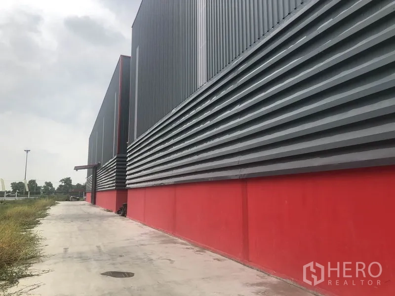 Warehouse for rent in Khlong Luang, Pathum Thani - Long side elevation of the warehouse featuring ventilated metal panels, service walkway and adjacent greenery.