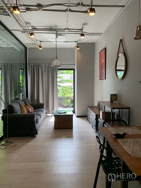 Condominium for sale in Watthana, Bangkok - Bright living room with grey sofa, pendant lights, light wood flooring, and glass door opening to a leafy balcony.