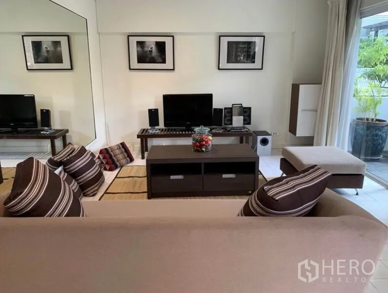 Townhouse for rent in Khlong Toei, Bangkok - Bright living room with beige sofa, striped cushions, media console and sliding glass door to a patio.