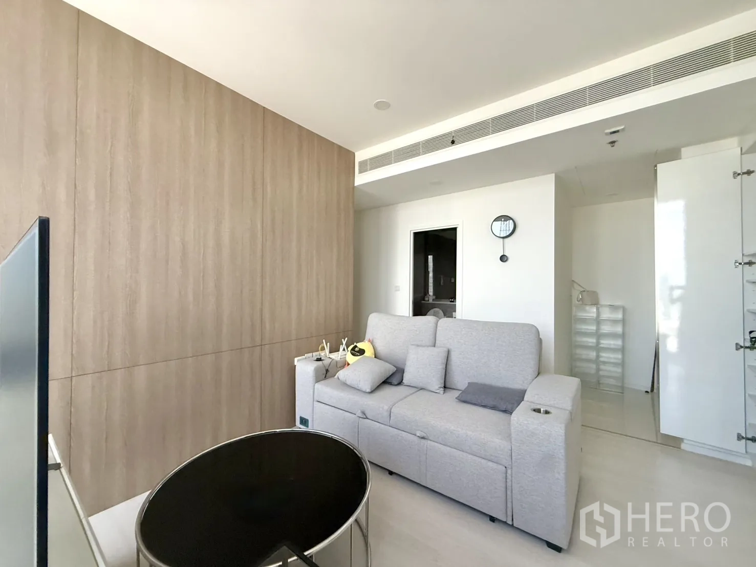 Condominium for sale in Chatuchak, Bangkok - Bright living area with grey sofa, wood accent wall, and doorway to bathroom.