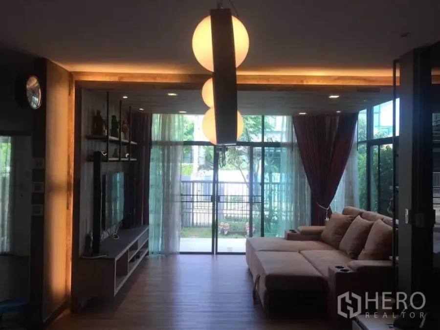 Detached House for sale in Pak Kret, Nonthaburi - Warm living room with floor-to-ceiling glass, pendant lights and an L-shaped sofa opening to the garden.