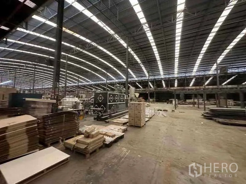 Factory for rent in Pathum Thani - Factory floor with pallets, machinery and crates beneath continuous skylights.