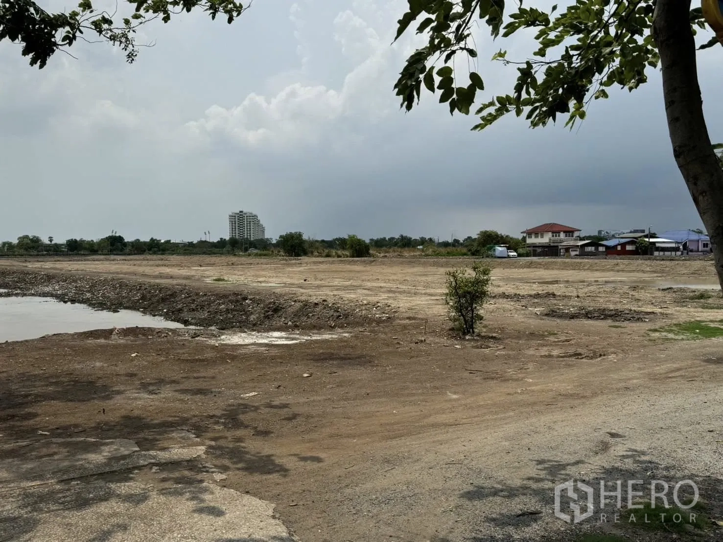 Land for sale in Bang Phli, Samut Prakan - Cleared land plot with a water feature, local homes, and distant mid-rise buildings under a cloudy sky.