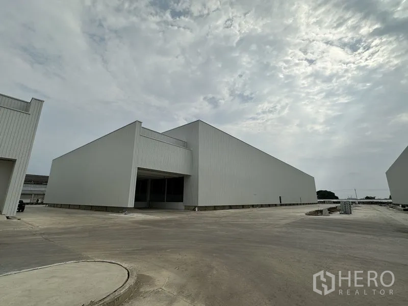 Factory for rent in Pathum Thani - New grey-clad factory buildings with a wide concrete driveway in an industrial park under a cloudy sky in Pathum Thani.