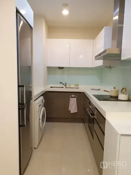 Condominium for rent in Pathum Wan, Bangkok - Modern U-shaped kitchen with glossy cabinets, hob, hood, fridge and in-unit washing machine.