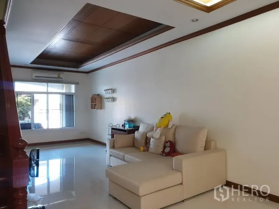 Townhouse for rent in Bang Kho Laem, Bangkok - Open-plan living room with L-shaped sofa, large window and recessed wooden ceiling.