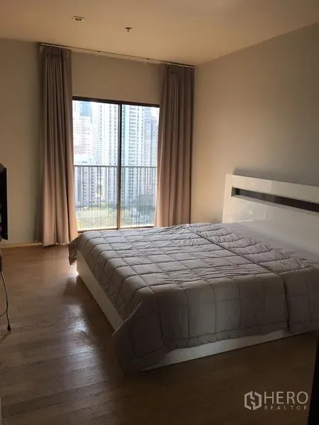 คอนโด ให้เช่าในBangkok - ห้องนอนแสงธรรมชาติ พื้นไม้ เตียงสีขาว และหน้าต่างบานใหญ่พร้อมระเบียงวิวเมือง