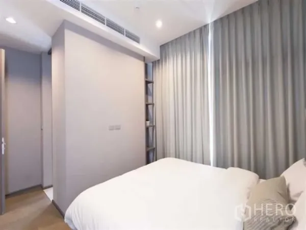 Condominium for rent in Bang Rak, Bangkok - Bedroom angle showing floor-to-ceiling curtains and a slim built-in shelving nook beside the doorway.