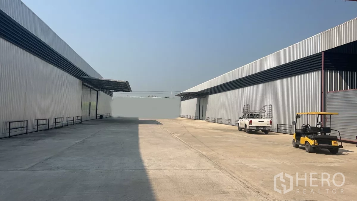 Warehouse for rent in Lat Lum Kaeo, Pathum Thani - Wide concrete yard inside a gated industrial compound between metal‑clad warehouses with parked vehicles and covered loading canopies.