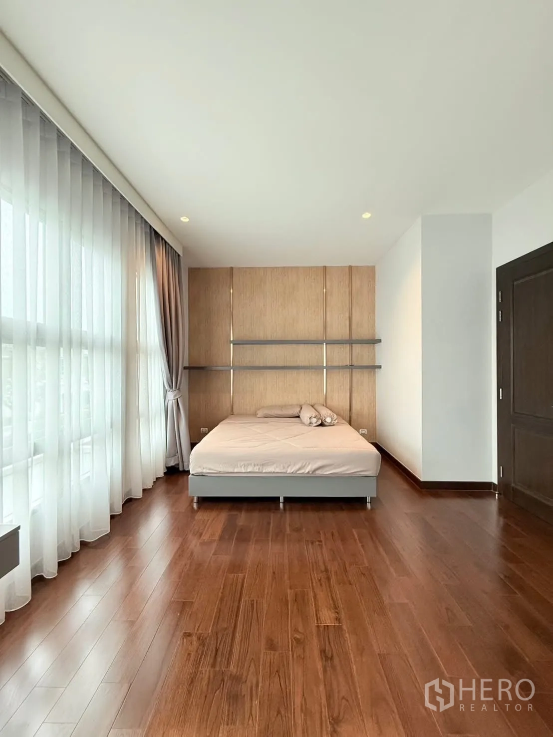 Detached House for rent or sale in Bang Phli, Samut Prakan - Spacious bedroom view with large windows, wood flooring, minimalist bed and panel feature wall.
