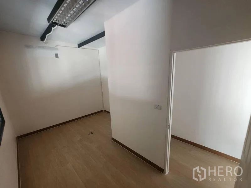 Warehouse for rent in Suan Luang, Bangkok - Compact office nook with fluorescent lighting and a connecting doorway.