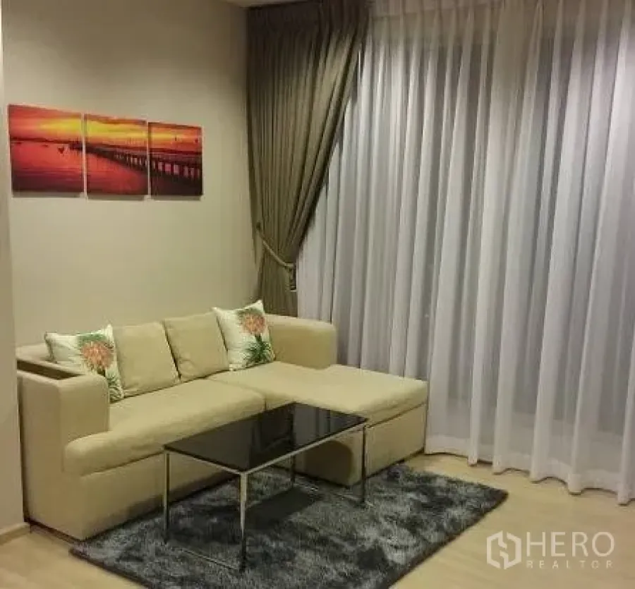 Condominium for rent in Sathon, Bangkok - Cozy living room with beige L-shaped sofa, glass coffee table and floor-to-ceiling sheer curtains.