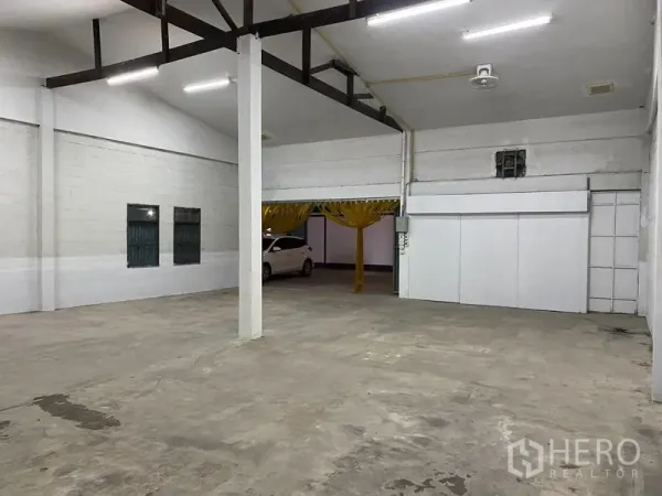 Warehouse for rent - Open-plan warehouse interior with concrete floor, white block walls, steel beams, bright strip lighting and a yellow PVC curtain entry with a parking bay beyond.