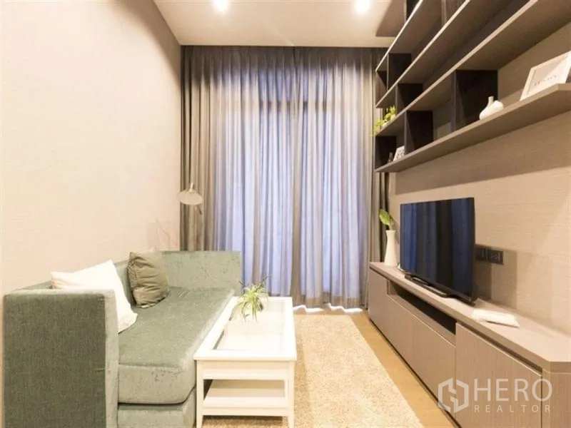 Condominium for rent in Bang Rak, Bangkok - Living space with long sofa, white coffee table, tall curtains and a media wall with shelving.