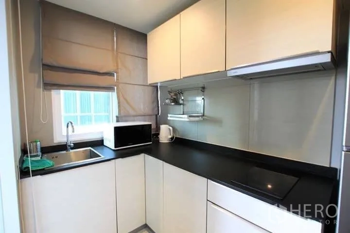 Condominium for rent in Watthana, Bangkok - Modern L-shaped kitchen with black countertop, sink, microwave and induction hob.