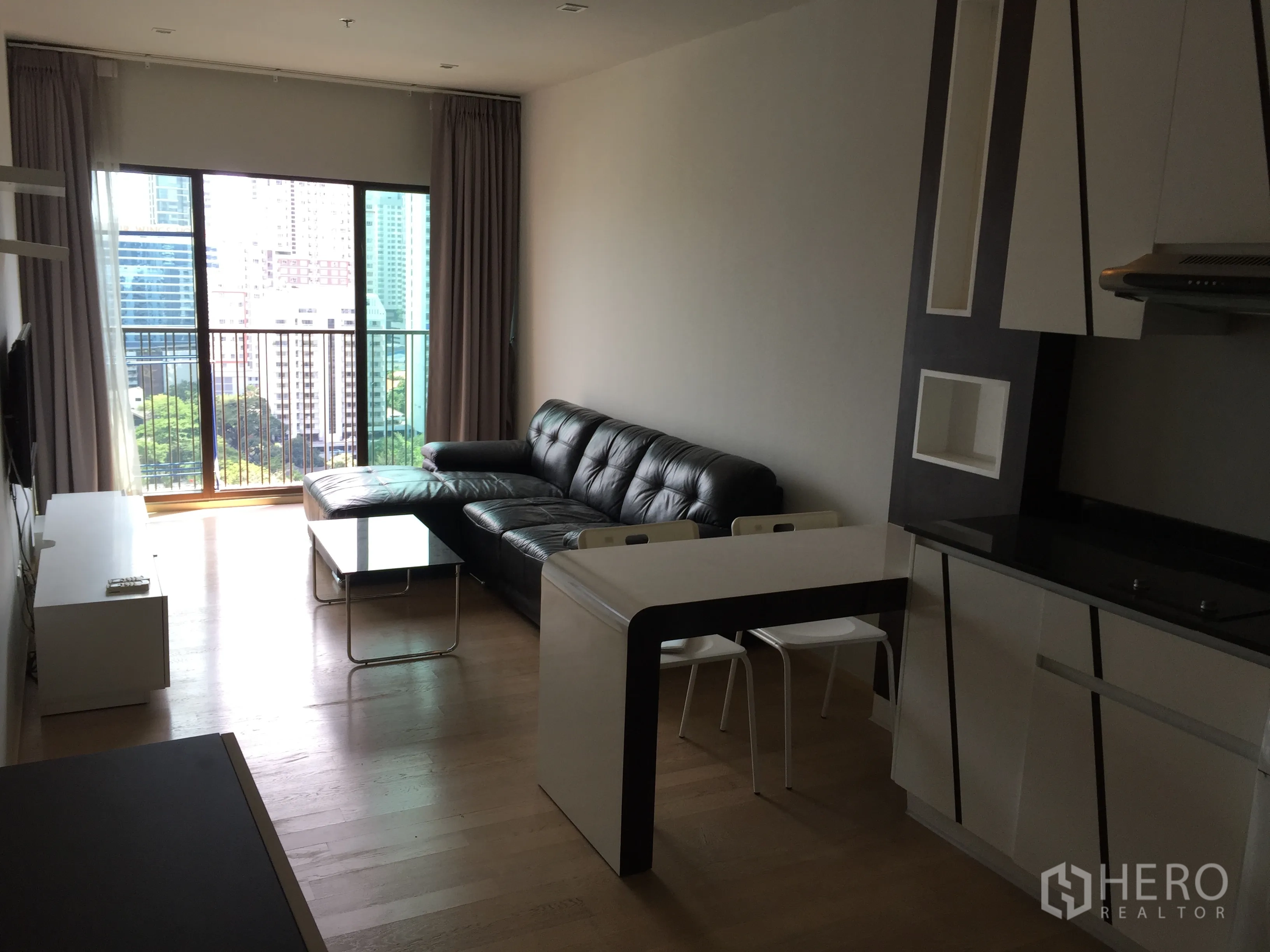 คอนโด ให้เช่าในBangkok - ห้องนั่งเล่นแบบเปิดโล่ง โซฟาหนังสีดำ ระเบียงชมวิวเมือง และครัวบิวท์อินกะทัดรัด.