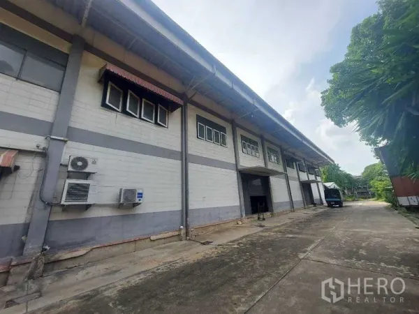 Warehouse for rent in Min Buri, Bangkok - Warehouse exterior with loading bay and roller shutter, high eaves and a wide driveway bordered by greenery.