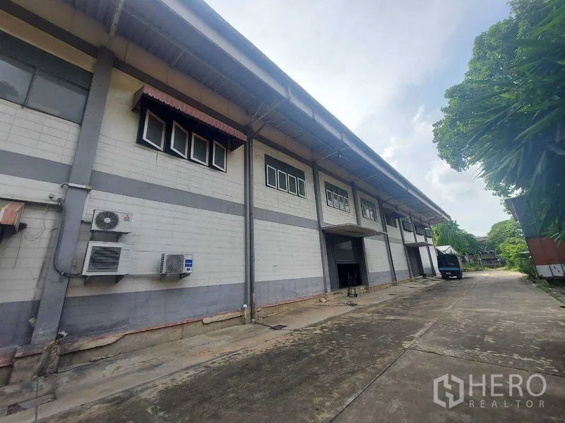 Warehouse for rent in Min Buri, Bangkok - Warehouse exterior with loading bay and roller shutter, high eaves and a wide driveway bordered by greenery.