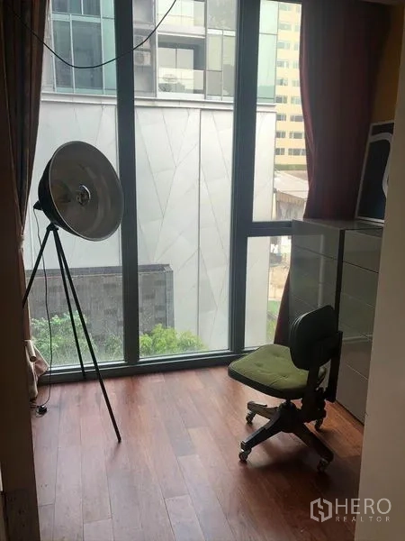 Condominium for sale in Watthana, Bangkok - Bright corner with floor‑to‑ceiling window, hardwood floor, studio lamp and chair overlooking city buildings.