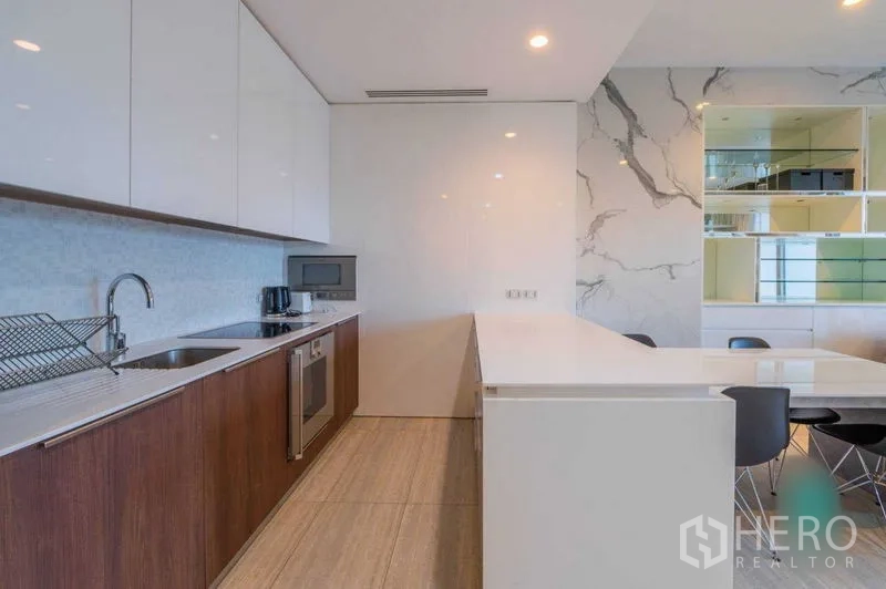 Condominium for rent in Pathum Wan, Bangkok - Sleek kitchen with white upper cabinets, wood base units, built-in oven, and a long quartz island beside a marble accent wall.