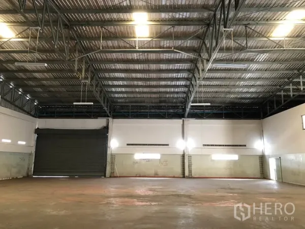 工厂 出租在Nonthaburi - 仓库内部空间开阔，钢桁架高挑屋顶、照明与卷帘门。