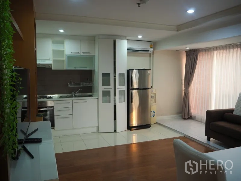 Condominium for rent or sale in Watthana, Bangkok - Open-plan kitchen with white cabinetry, stainless refrigerator, and large curtained window.