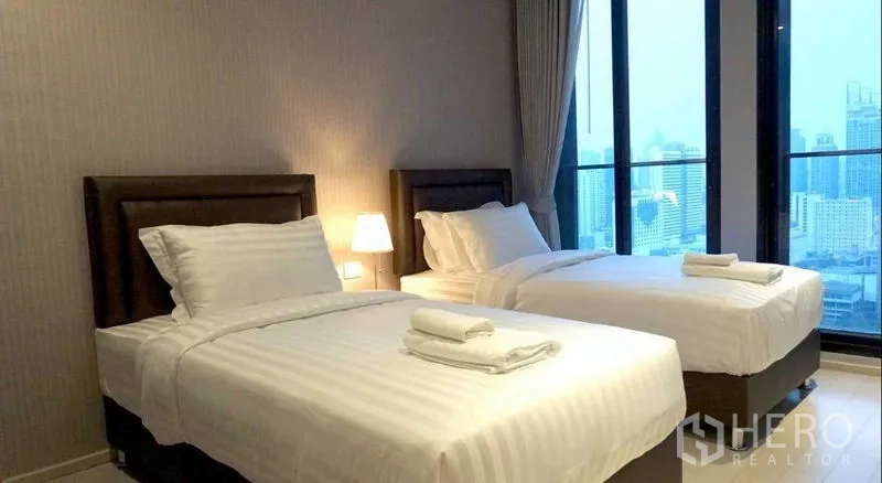 Condominium for rent in Pathum Wan, Bangkok - Twin bedroom with bedside lamp and floor-to-ceiling windows overlooking the city skyline.