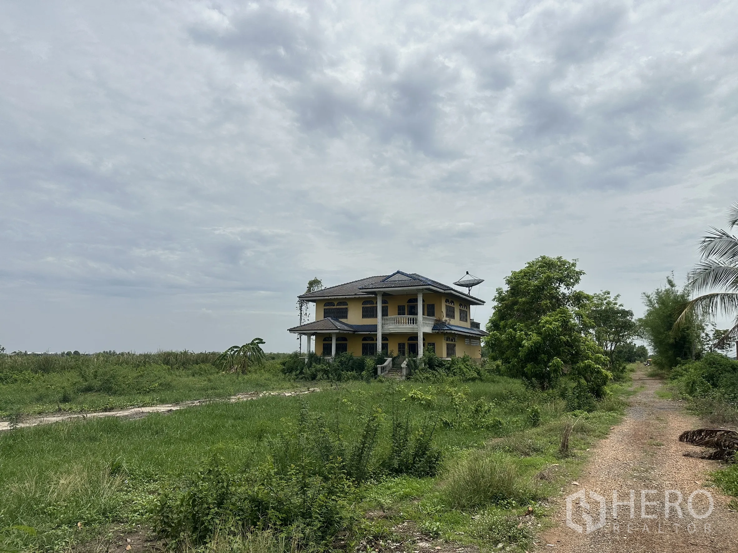 Land for sale in Bang Nam Priao, Chachoengsao - Two-storey rural house set within a large grassy field beside a dirt track.