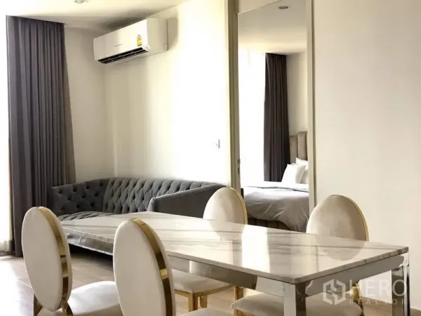 Condominium for rent or sale in Watthana, Bangkok - Open‑plan dining with marble table and tufted gray sofa, plus a sliding doorway leading into a light‑filled bedroom.