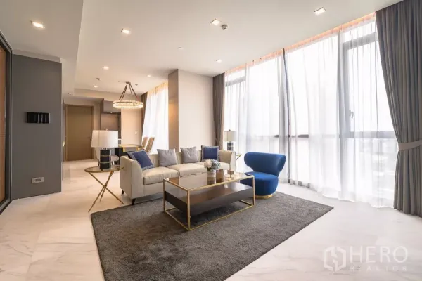 公寓 出租在Watthana, Bangkok - 采光充足的客厅，落地纱帘、米色沙发、蓝色单椅、金属茶几与吊灯。