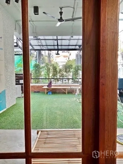 อาคารพาณิชย์ ให้เช่าในPathum Wan, Bangkok - มองผ่านกรอบไม้ไปยังพื้นที่กึ่งเอาท์ดอร์ พื้นหญ้าเทียม พัดลมเพดาน และแนวต้นไม้