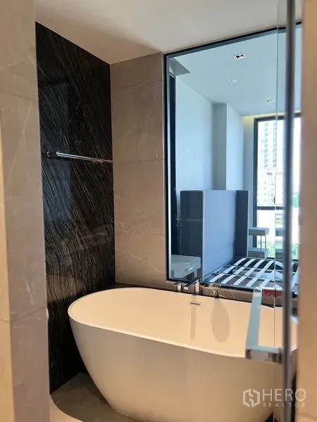 公寓 出售在Watthana, Bangkok - 当代浴室，独立式椭圆浴缸，岩纹装饰墙，大窗望向卧室与城市景观。